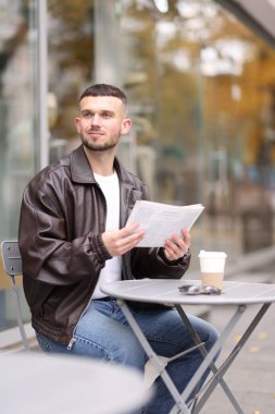 Genç adam açık kafedeki masada gazete okuyor.