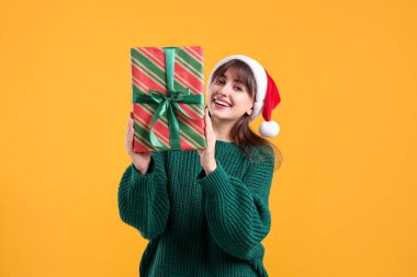 Sarı arka planda hediye kutusuyla Noel Baba şapkalı gülümseyen kadın. Noel ve Yeni Yıl Kutlaması