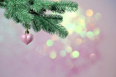 Köknar dalından sarkan kalp şeklinde bir Noel süsü pembe arka plandaki bulanık ışıklara karşı