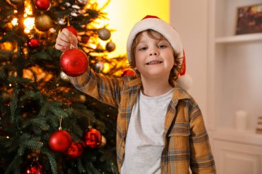 Noel Baba şapkalı küçük çocuk evde Noel ağacı süslüyor.