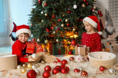 Noel Baba şapkalı şirin çocuklar ve evde Noel ağacının yanında süsleri var.