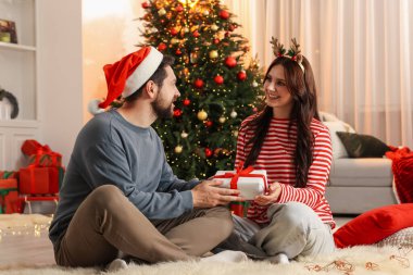 Hediye kutusu ve komik şapkalı mutlu çift Noel için dekore edilmiş odada birlikte vakit geçiriyorlar.
