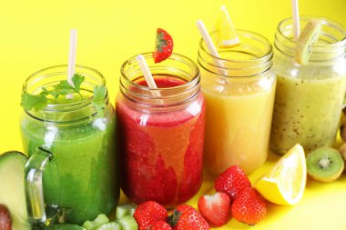 Mason kavanozlarındaki farklı lezzetli içecekler ve sarı arka plandaki malzemeler, yakın plan.