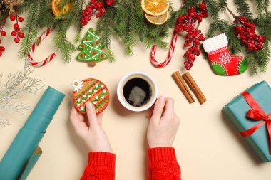 Bir fincan aromatik kahve, noel kurabiyesi, köknar ağacı dalları, bej arka planda hediye kutusu ve dekoru olan bir kadın, üst görünüm.