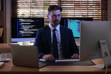 Takım elbiseli ve gözlüklü programcı ofisteki ahşap masada bilgisayarla çalışıyor.