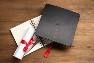 Mezuniyet şapkası, diploma, defter ve kalem ahşap masanın üstünde.