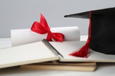 Mezuniyet şapkası, diploma ve beyaz ahşap masa üzerindeki defterler arka planda, yakın plan.