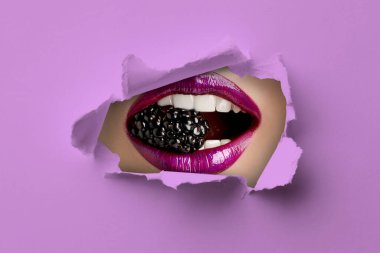 Ağzında böğürtlen tutan genç bir kadın, yakın plan. Menekşe kağıdındaki yırtık delikten dudaklarına bak.