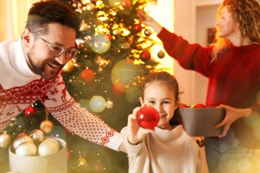 Mutlu yıllar 2026 tebrik kartı, Bokeh ışıkları. Mücevherli küçük bir kız ve ailesi evde Noel ağacı süslüyorlar.