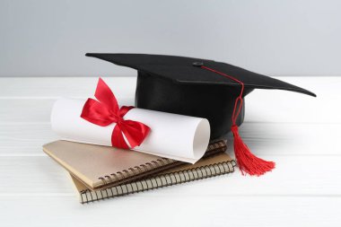 Mezuniyet şapkası, diploma ve beyaz ahşap masa üzerindeki defterler.