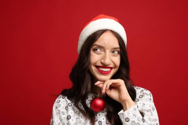 Çekici bir kadın Noel Baba şapkası takıyor ve kırmızı arka planda Noel ağacı süsü var.