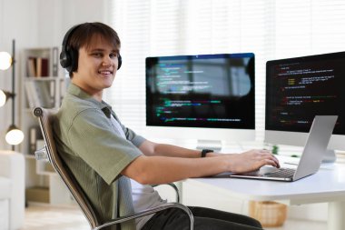 Ofisteki beyaz masada dizüstü bilgisayarla çalışan kulaklıklı gülen programcı.