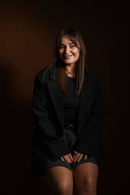 Koyu kahverengi arka planda, sandalyede oturan şık elbiseli güzel bir genç kadın.