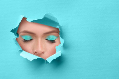 Parlak makyajlı genç bir kadın, yakın plan. Açık mavi kağıttaki yırtık delikten gözlere bak