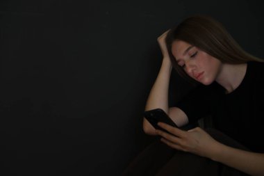 Kapalı alanda siyah duvarın yanında akıllı telefonu olan üzgün genç kız, mesaj için yer var.