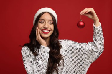 Çekici bir kadın Noel Baba şapkası takıyor ve kırmızı arka planda Noel ağacı süsü var.