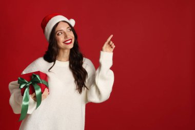 Noel Baba şapkalı ve Noel hediyeli güzel bir kadın kırmızı arka plandaki bir şeyi işaret ediyor. Metin için boşluk