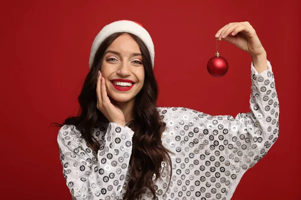 Çekici bir kadın Noel Baba şapkası takıyor ve kırmızı arka planda Noel ağacı süsü var.