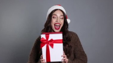 Heyecanlı kadın gri arka planda Noel hediyesi olan Noel Baba şapkası giyiyor.