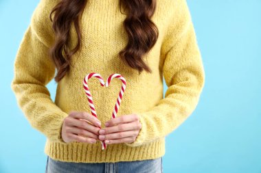 Sarı kazaklı kadın açık mavi arka planda baston şekerle kalp yapıyor, yakın plan. Mutlu Noeller ve mutlu yıllar.