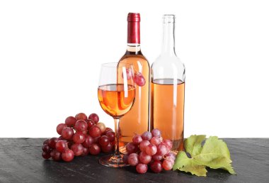 Bardakta gül şarabı, şişeler, üzümler ve yapraklar siyah masada beyaz arka planda