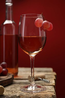 Cam, şişe, üzüm ve tirbuşonlu gül şarabı ahşap masada kırmızı arka planda, yakın planda.