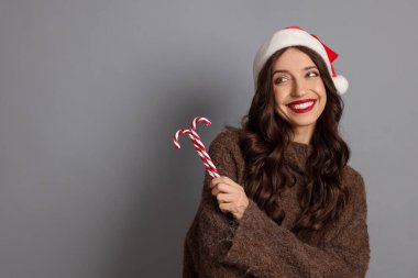 Noel Baba şapkası ve baston şekeriyle güzel bir kadın gri arka plan, mesaj için yer. Noel sezonu