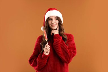 Bej arka planda lolipopu olan, Noel Baba şapkalı ve kazaklı düşünceli bir kadın. Mutlu Noeller ve mutlu yıllar.