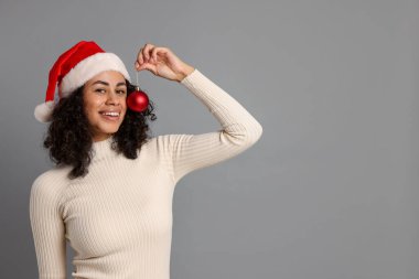 Gri arka planda Noel ağacı süsü olan Noel Baba şapkası takan çekici bir kadın. Metin için boşluk