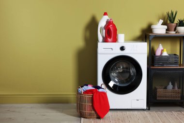 Modern çamaşır makinesi, deterjanlar ve zeytin duvarının yanındaki giysiler.