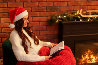 Noel Baba şapkalı kadın şöminenin yanında kitap okuyor.