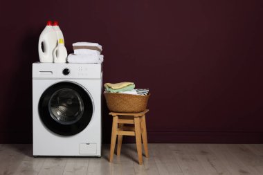 Modern çamaşır makinesi, deterjanlar ve bordo duvarın yanındaki giysiler.