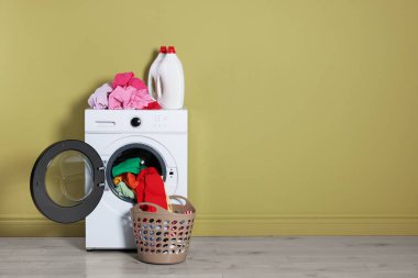 Modern çamaşır makinesi, deterjanlar ve zeytin duvarının yanındaki giysiler.