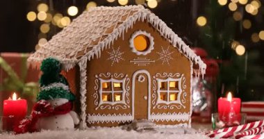 Dekoratif kar, zencefilli çöreklerin evine düşüyor ve masanın üzerindeki Noel dekoru bulanık ışıklara karşı, yakın plan.