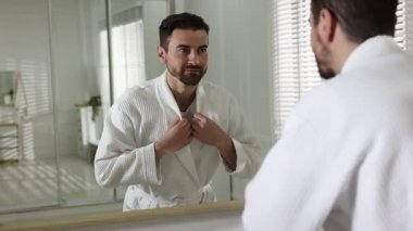 Beyaz bornoz giyen yakışıklı adam aynada kendine bakıyor.