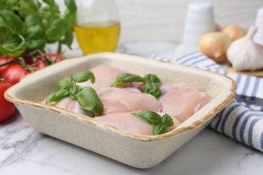 Çiğ tavuk butları fırın kabında fesleğen ve beyaz mermer masada ürünler, yakın plan.