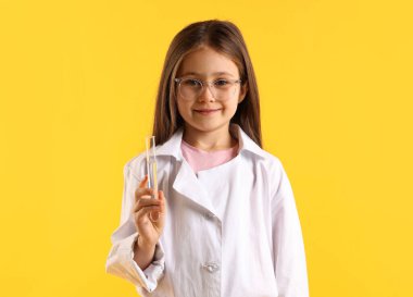 Laboratuvar önlüğü ve sarı arka planda test tüpü olan gülümseyen küçük kız portresi. Çocuk ve bilim