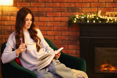 Kakao içen kadın şöminenin yanında kitap okuyor.