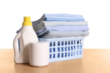 Temiz elbiseli çamaşır sepeti ve ahşap masadaki deterjanlar beyaz arka planda.
