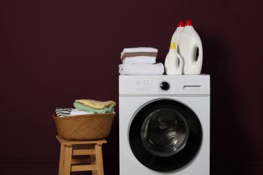 Modern çamaşır makinesi, deterjanlar ve bordo duvarın yanındaki kıyafetler.