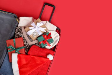 Noel tatili. İçinde Noel Baba şapkası, kıyafetler ve kırmızı arka planda hediye kutuları olan bavulu aç. Metin için boşluk