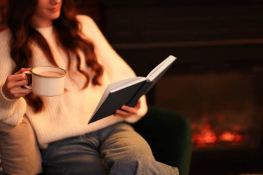 Kakao içen kadın şöminenin yanında kitap okuyor, yakın plan. Metin için boşluk
