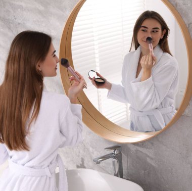 Bornozlu güzel bir kadın aynanın yanında makyaj yapıyor.