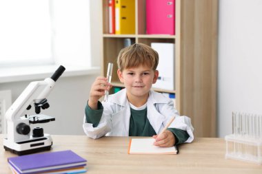 Elinde test tüpü ve mikroskop olan gülümseyen küçük çocuk içeride masa başında yazıyor. Çocuk ve bilim
