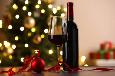 Bir kadeh kırmızı şarap, şişe ve Noel süsü ahşap masada arka planda bulanık ışıklarla.