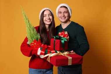 Şapkalı mutlu bir çift, sarı arka planda bir Noel ağacı ve hediyeleri.