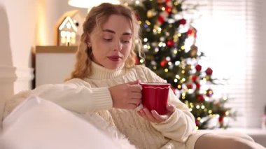 Kanepede bir fincan sıcak içecekle gülümseyen kadın Noel için dekore edilmiş odada yanıp sönen ışıklara karşı.