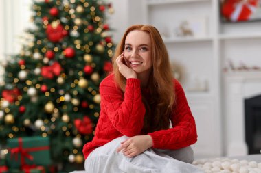 Evdeki bulanık ışıklara karşı kanepede süveter giymiş gülümseyen kadın portresi. Mutlu Noeller ve mutlu yıllar.