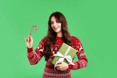 Yeşil arka planda şeker kamışı ve hediye kutusuyla süveter giyen gülümseyen kadın. Mutlu Noeller ve mutlu yıllar.