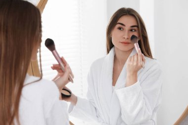 Bornozlu güzel bir kadın aynanın yanında makyaj yapıyor.